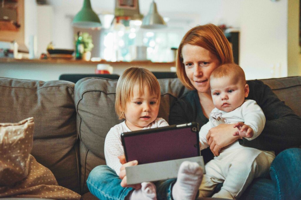 Kuvassa äiti ja kaksi pientä lasta istumassa sohvalla. Toinen on noin vuoden ikäinen ja toinen noin kolmivuotias. Lapset istuvat äidin sylissä ja he kaikki katsovat jotain ohjelmaa tai sisältöä tabletilta.